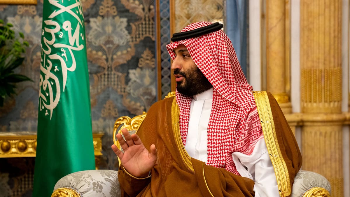 Saudi Crown Prince Mohammed bin Salman in a red-checked headdress and brown robe, gesturing beside the green Saudi flag