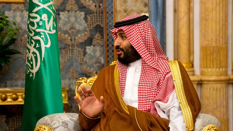 Saudi Crown Prince Mohammed bin Salman in a red-checked headdress and brown robe, gesturing beside the green Saudi flag