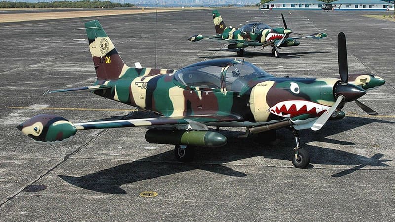 Two camouflaged SF-260 propeller aircraft with shark-mouth markings parked on an airfield apron
