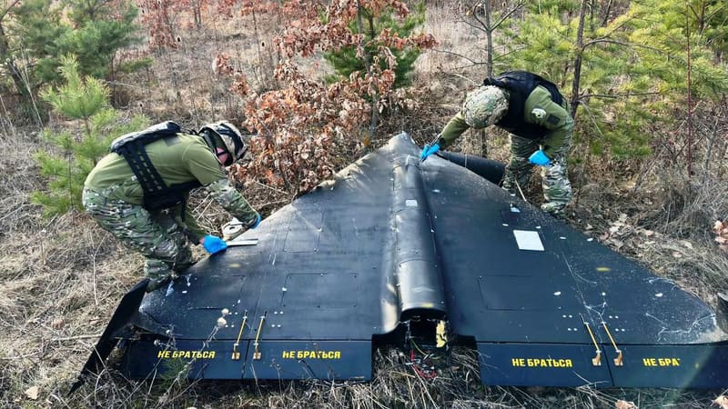Black delta-wing Shahed-type drone lying in a forest as two Ukrainian police bomb-disposal officers examine it