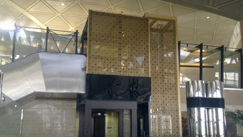 Interior of an airport terminal with a gold perforated screen, a stainless-steel escalator and a triangular-grid ceiling