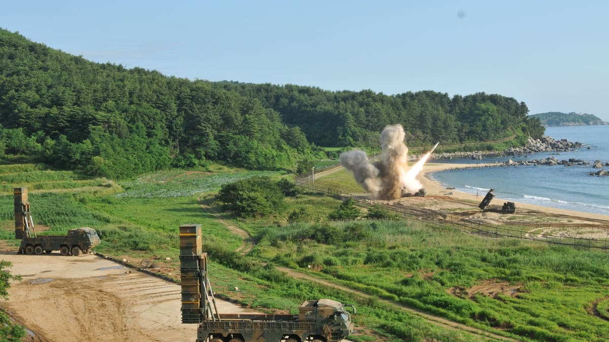 Two South Korean missile launch trucks with raised canisters on a coastal field as a U.S. rocket launcher fires on the beach