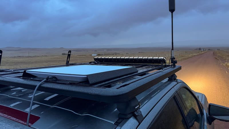 A flat Starlink terminal on a vehicle roof rack under a stormy sky, with a whip antenna and a dirt road beside the car