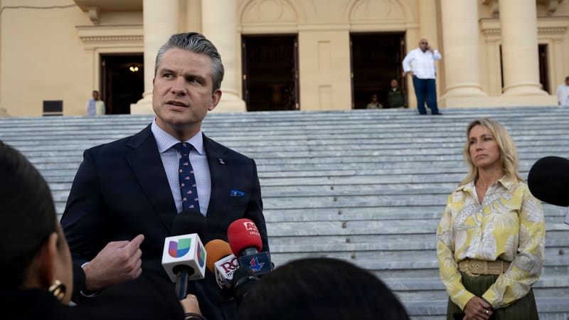 Pete Hegseth in a dark suit speaks into a cluster of press microphones outside a columned palace