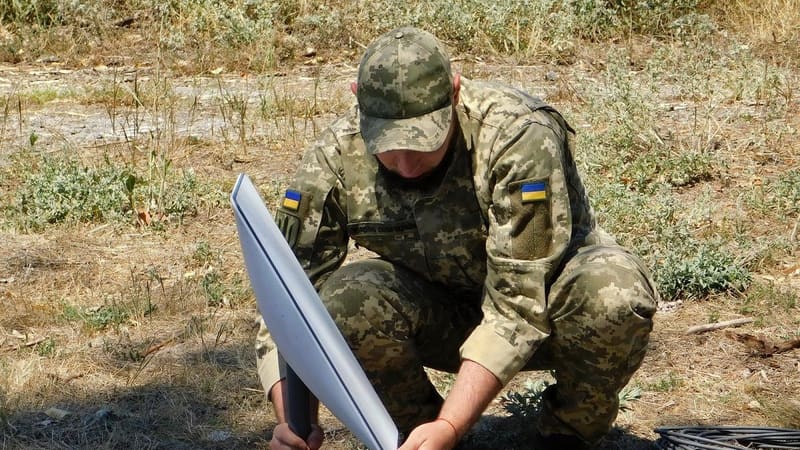A Ukrainian soldier in camouflage crouches to set up a white Starlink satellite dish on a tripod in a dry field