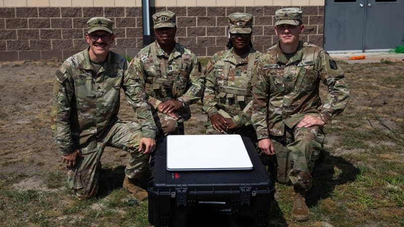 Four U.S. Army Reserve soldiers kneel behind a flat white Starshield satellite terminal resting on a black case