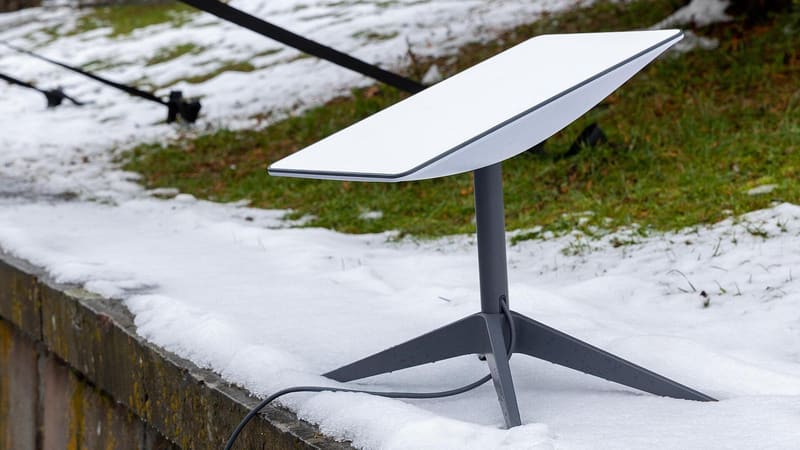 A white Starlink satellite dish on a grey tripod stands on a snow-covered stone wall
