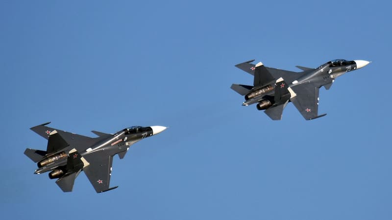 Two dark grey Russian Navy Su-30SM fighters with side numbers 77 and 75 flying against a clear blue sky