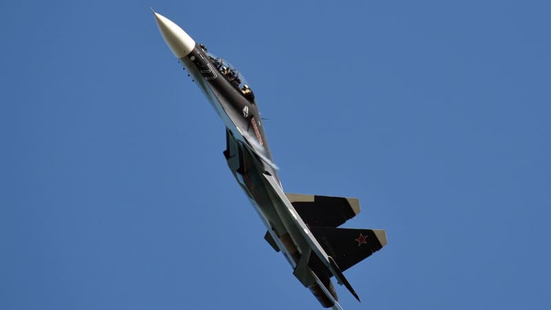 A dark grey Russian Navy Su-30SM fighter with side number 77 climbing steeply against a blue sky