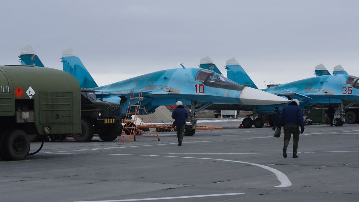 Pilots walk across an apron past parked blue Su-34 bombers numbered 10 and 35, with a fuel truck at left