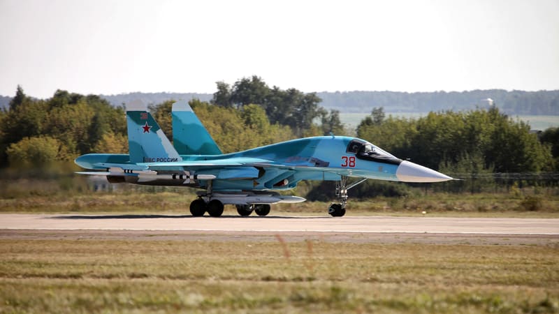 A blue camouflaged Su-34 fighter-bomber with red number 38 taxiing on a runway beside a grass strip
