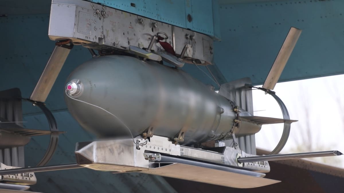 A grey FAB-500T bomb fitted with folding UMPK glide wings hangs under the wing of a blue Su-34