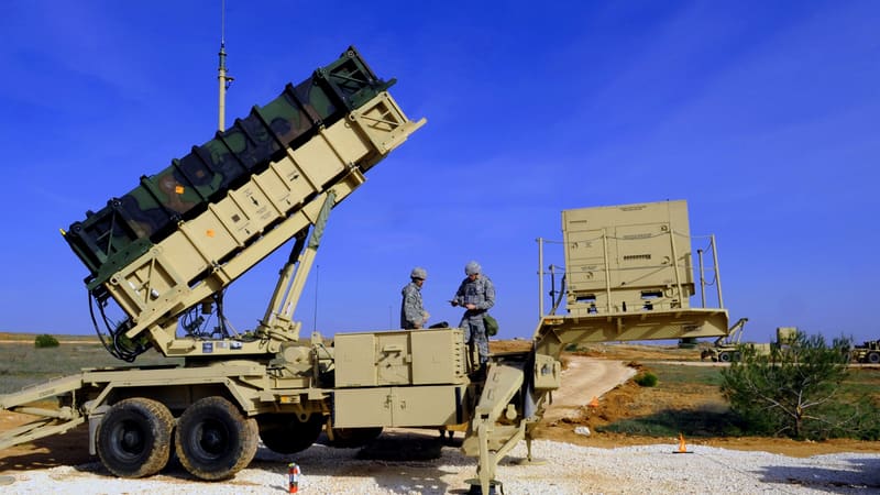 Two U.S. soldiers beside a raised Patriot missile launcher on a gravel site under a blue sky
