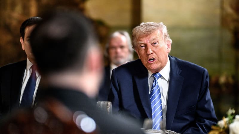 Donald Trump in a navy suit and blue tie speaks across a formal table; a blurred figure is seen from behind