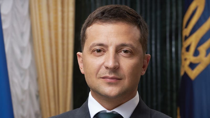 Official portrait of Ukrainian President Volodymyr Zelensky in a dark suit, with a flag and standard behind him.