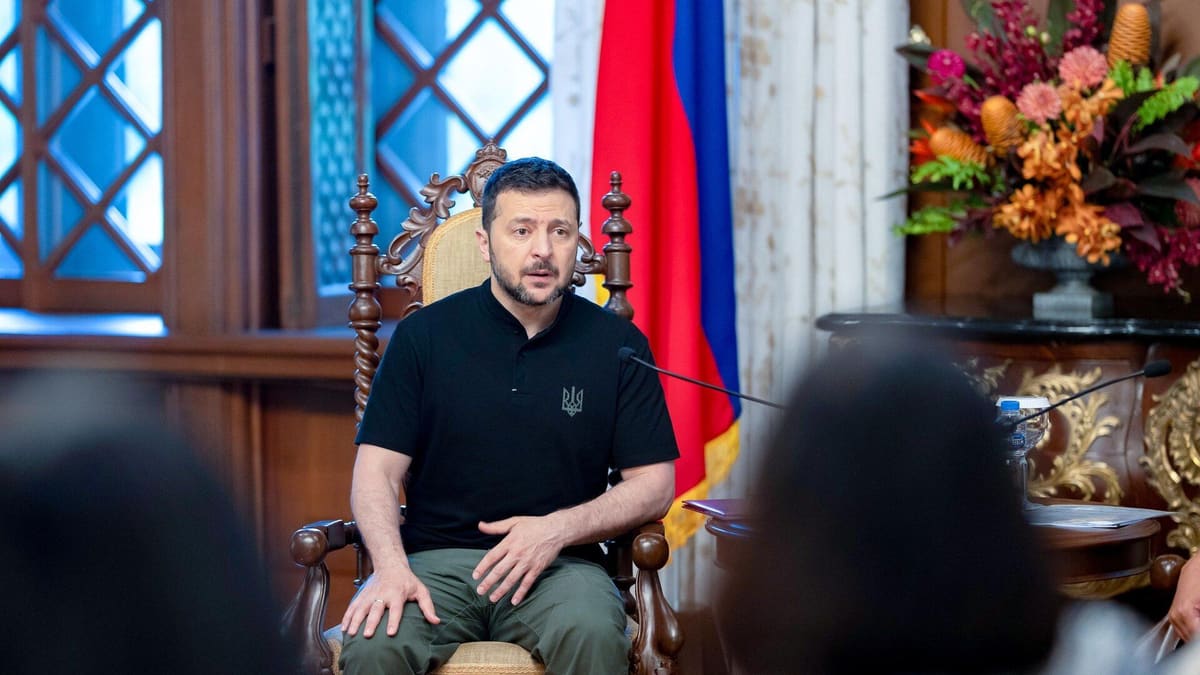 Zelensky in a black shirt with a trident emblem sits in a carved wooden chair, speaking, with a flag and flowers behind him.