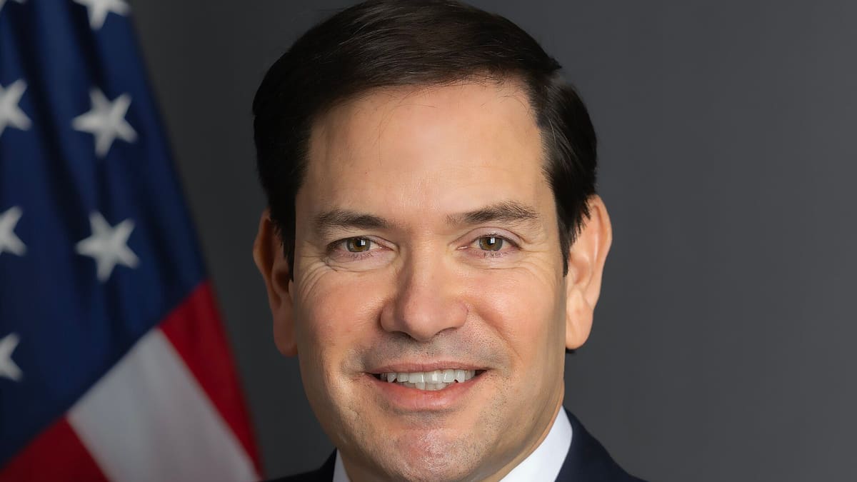 Marco Rubio in a dark suit smiling in front of a US flag