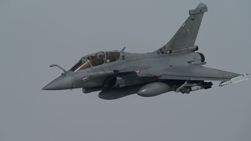 A grey two-seat French Rafale B with guided bombs and fuel tanks under its wings flying against a hazy grey sky