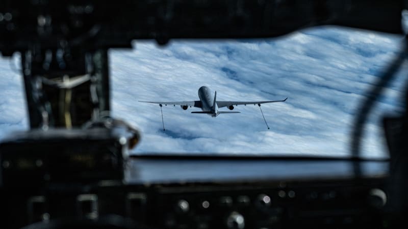 A French A330 MRTT tanker trailing two refuelling hoses from its wing pods, seen over clouds through a KC-135 cockpit window