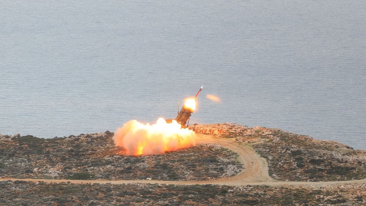 A Patriot missile rises from a launcher on a rocky headland above the sea amid a bright orange fireball.