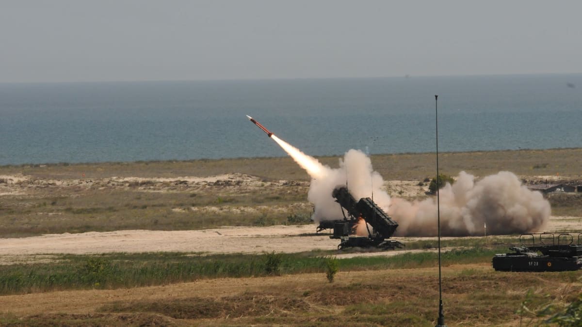 A U.S. Army Patriot launcher fires an interceptor beside a second launcher on a grassy coastal range above the sea.