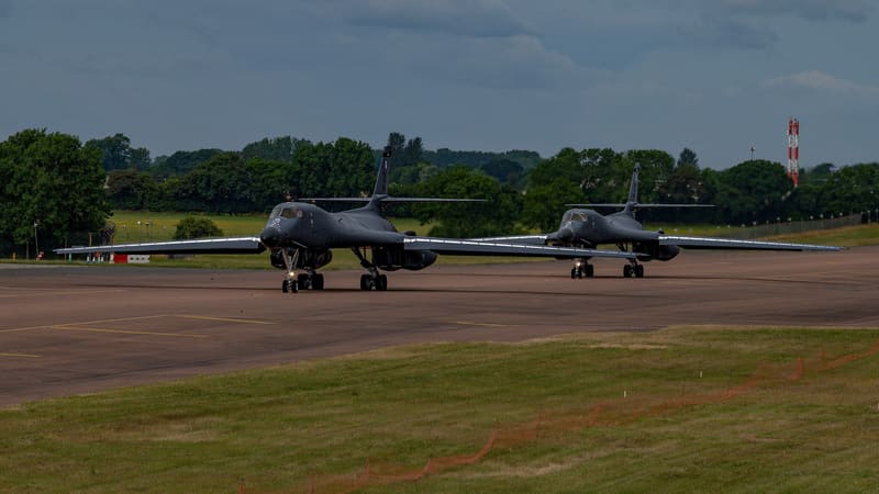 Two dark grey B-1B Lancer bombers with wings spread taxiing one behind the other on an airfield under a cloudy sky