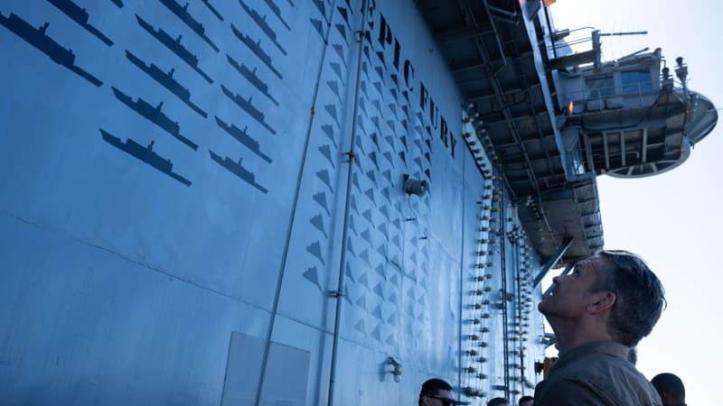 Pete Hegseth looks up at rows of ship and drone silhouettes painted on USS Abraham Lincoln's island under the words Epic Fury