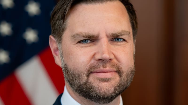 Official portrait of US Vice President JD Vance, a bearded man, with a blurred American flag behind him