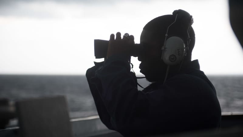 A US Navy lookout in silhouette raises binoculars on the bridge wing of a destroyer