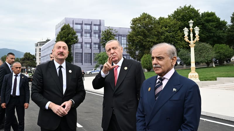 Erdoğan, gesturing with one hand, stands between Ilham Aliyev and Shehbaz Sharif on a paved road