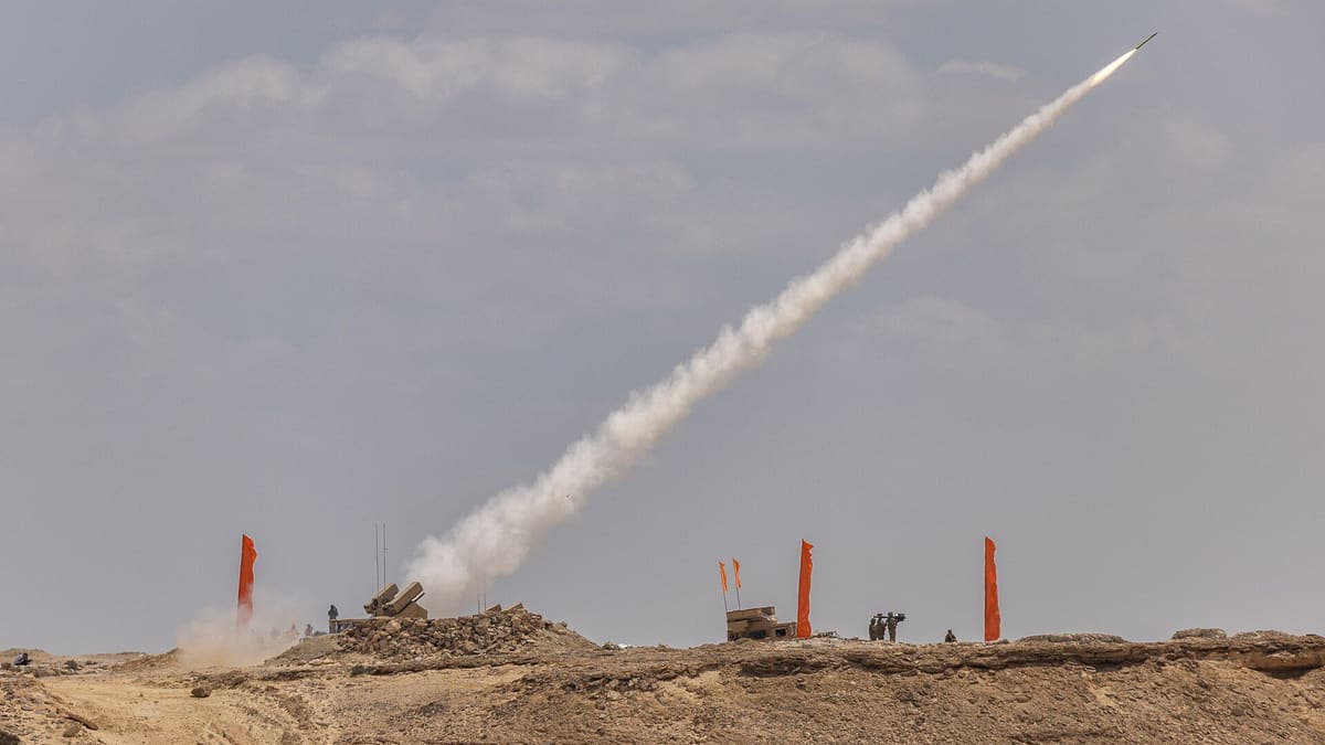 A short-range air defence missile streaks into a grey sky from a launcher on a desert ridge marked with orange flags.