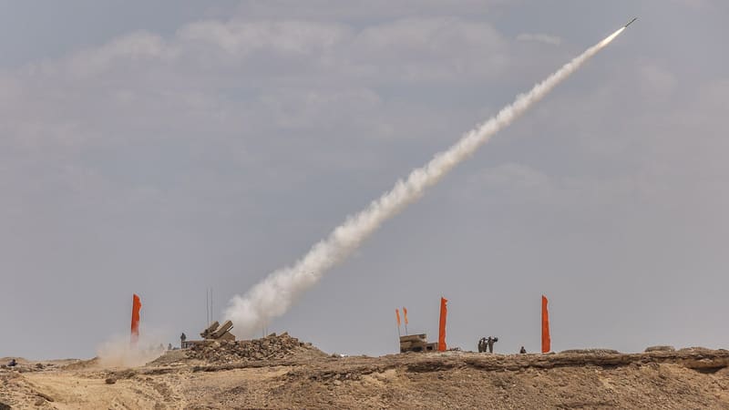 A short-range air defence missile streaks into a grey sky from a launcher on a desert ridge marked with orange flags.