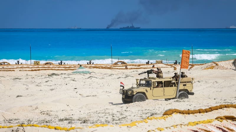 Egyptian soldiers in a tan Humvee with a mounted machine gun on a beach, warships and smoke offshore