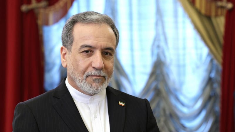 Abbas Araghchi in a dark pinstripe suit with an Iranian flag pin, in front of red and white curtains
