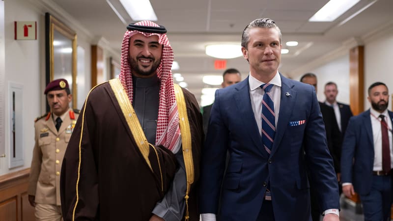 Khalid bin Salman in a red-and-white headdress and gold-trimmed brown robe walks beside Pete Hegseth down a corridor