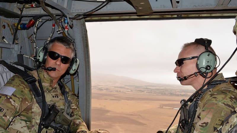 Two U.S. Army officers wearing headsets and sunglasses sit in an open helicopter door above a desert plain