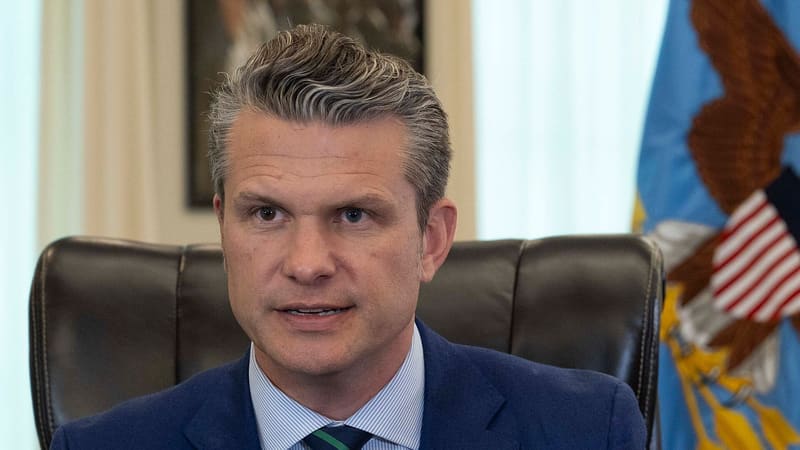 Pete Hegseth in a blue suit speaks and points from a leather chair beside a Defense Department flag