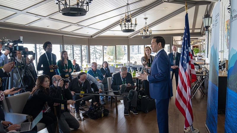 Marco Rubio in a navy suit speaks to seated and kneeling journalists beside a US flag.