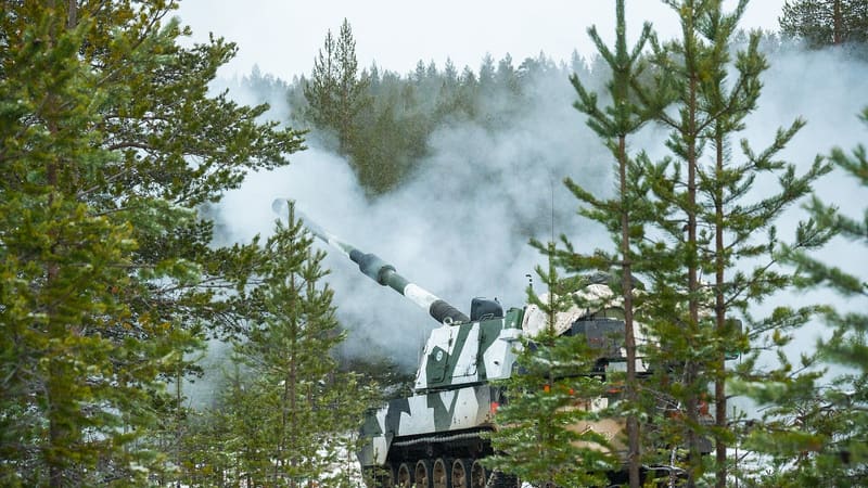 A Finnish Army K9 Moukari tracked self-propelled howitzer fires among pine trees, smoke rising behind the raised barrel