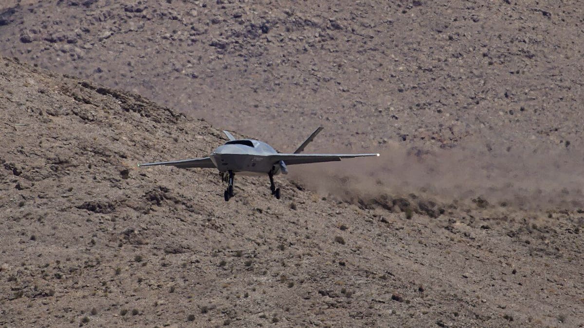 A grey YFQ-42A drone with its landing gear down flies low past a rocky desert hillside